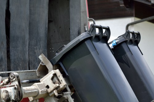 The image shows two large black wheelie bins with closed lids, positioned side by side on a pavement or driveway area. The bins are made of plastic with a smooth, slightly reflective surface, and are situated against a grey wooden fence or wall. To the left, part of a metal structure, possibly a waste collection truck or bin lorry, is visible, with mechanical components and a gripping mechanism for lifting the bins. The environment appears to be an outdoor setting in a residential or suburban location, with natural daylight illuminating the scene. The bins are typical of rubbish or waste collection containers used in areas such as Twickenham. This setup suggests a rubbish collection or waste removal process, relevant to the services offered by Garden Clearance Twickenham.