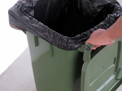 A close-up view of a green plastic wheelie bin with a black plastic rubbish bag inside, partially folded over the rim. A person's hand is gripping the bin's handle on the right side, preparing for rubbish removal. The bin is positioned on a grey concrete surface, with the background showing a plain white or neutral-colored surface, likely an outdoor or driveway setting in Twickenham. The image emphasizes the practical use of a wheelie bin for waste collection or clearance services, consistent with professional rubbish removal activities that a company like Garden Clearance Twickenham might undertake in the local area.