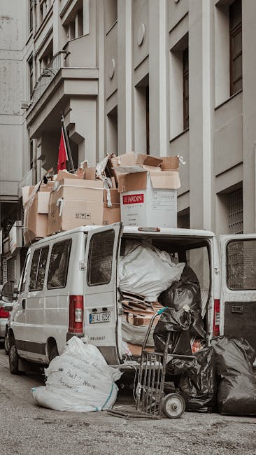 A small white flatbed truck is parked on an urban street, overloaded with three large, transparent plastic bags filled with mixed waste plastic bottles, containers, and packaging. The bags are secured