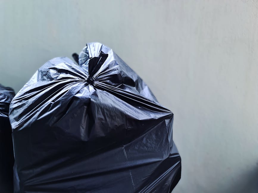Close-up view of several black plastic rubbish bags, which are tightly knotted at the top, resting against a plain light-colored wall. The bags are made of shiny, crinkled plastic material, reflecting