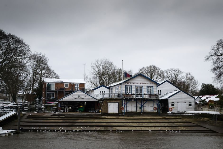 A lakeside building labeled 'Vikenham Rowing Club' situated on a narrow strip of land along a waterway, featuring a predominantly white exterior with dark blue trim and window frames. The structure in